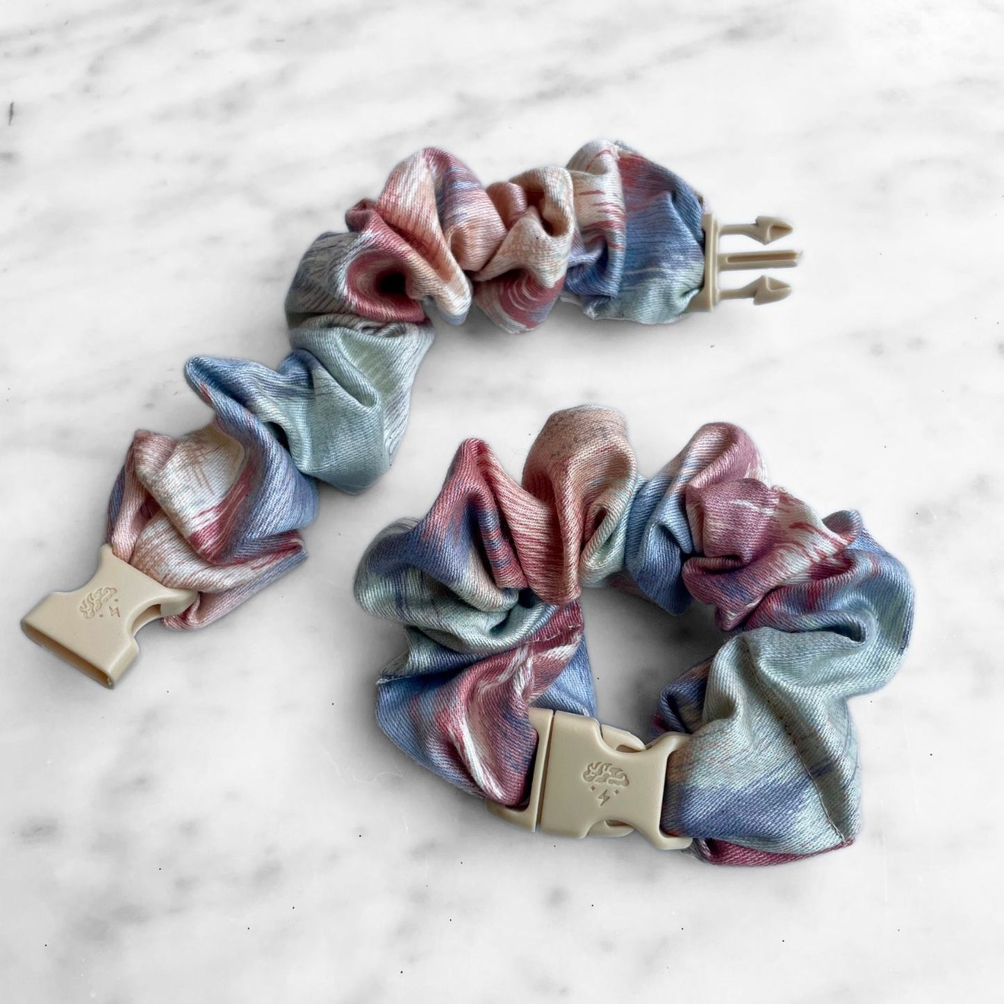 Two pink and blue watercolor Buckle Scrunchies on white marble, one open with the buckle unclipped
