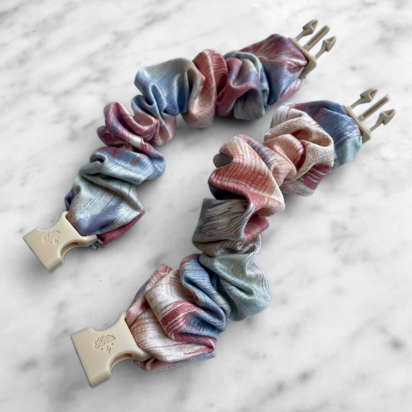 Two pink and blue watercolor Buckle Scrunchies laid open on marble with tan buckles