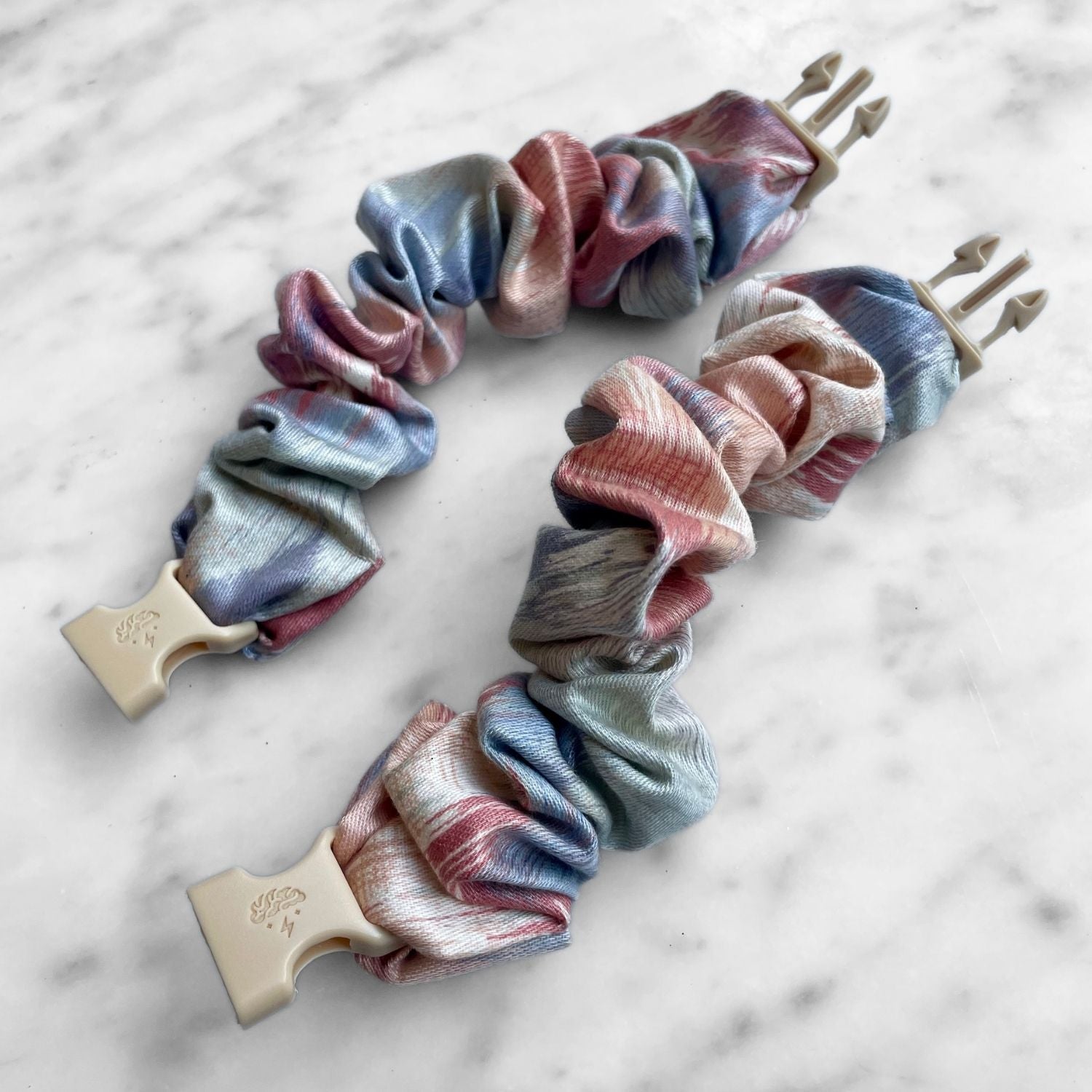Two pink and blue watercolor Buckle Scrunchies laid open on marble with tan buckles